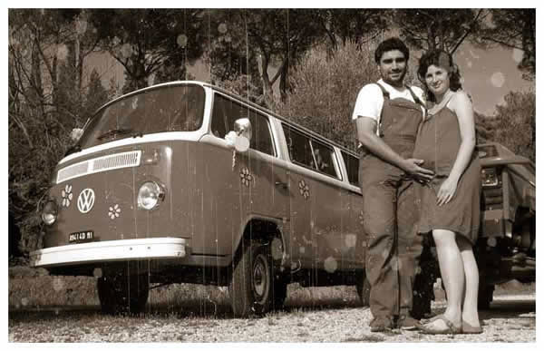 Una coppia sorridente si trova davanti a un furgone Volkswagen d'epoca decorato con adesivi floreali. L'uomo, in tuta, abbraccia la vita della donna. La foto ha un tono seppia che le conferisce un'atmosfera nostalgica e retrò.